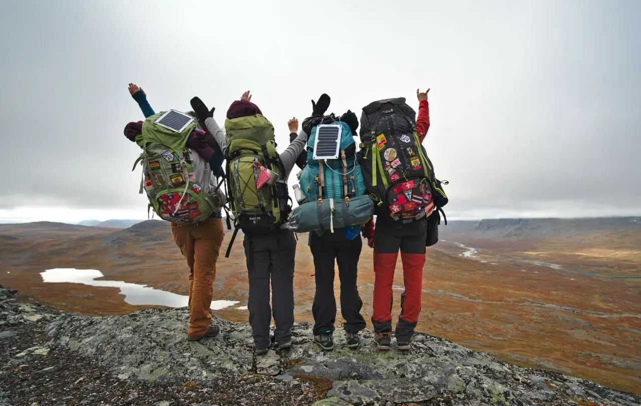 Quatre randonneurs, sacs &agrave; dos charg&eacute;s, bras lev&eacute;s, contemplent un paysage de toundra. Une aventure en course &agrave; &eacute;tapes, pleine de d&eacute;couvertes.