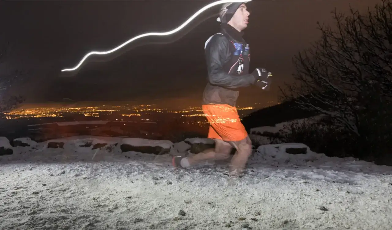 Un coureur Asics Sainte Lyon, en pleine nuit, traverse un paysage enneigé avec la ville illuminée en arrière-plan.