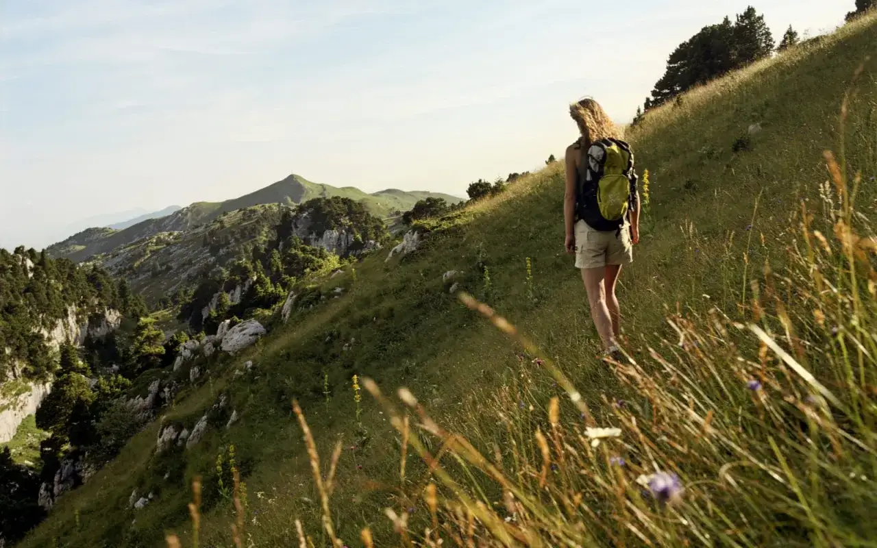 Une randonneuse profite d'un magnifique trek dans le Jura, avec des montagnes verdoyantes et un ciel bleu.