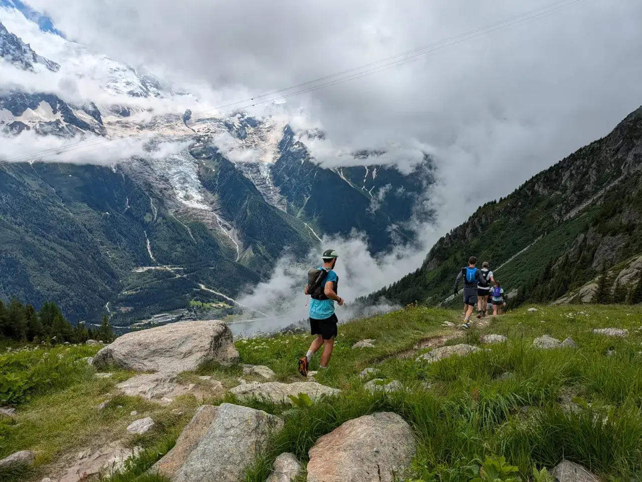 Coureurs en entraînement pour l'UTMB, traversant un sentier de montagne verdoyant avec des glaciers et des nuages en arrière-plan.