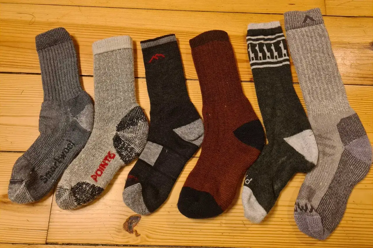 Six paires de chaussettes de randonnée, de différentes couleurs et motifs, sont alignées sur un plancher en bois. Quelle chaussette pour randonnée choisir ?
