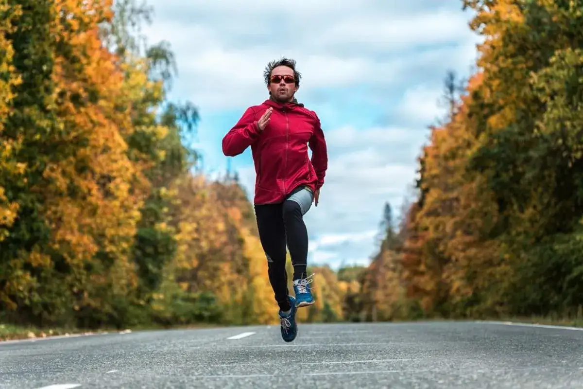 Un homme court sur une route bord&eacute;e d'arbres aux couleurs d'automne. Il porte une veste rouge et des lunettes de soleil. Son entra&icirc;nement est un exemple fractionn&eacute; court.
