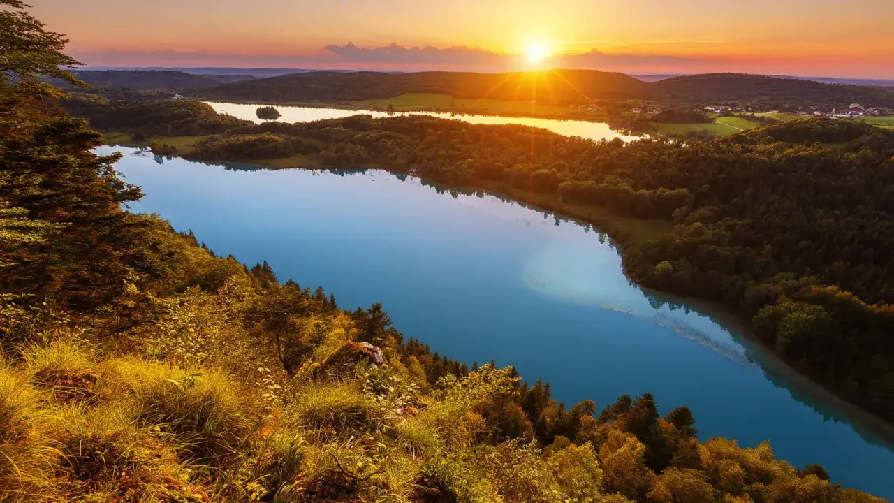 Vue panoramique d'un lac bleu intense au coucher du soleil, parfait pour une randonn&eacute;e Jura 2 jours. For&ecirc;ts verdoyantes et collines en arri&egrave;re-plan.