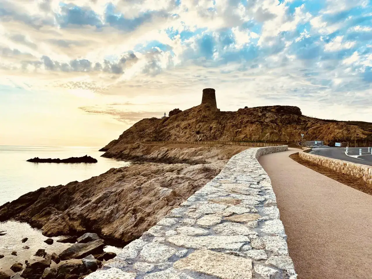 Chemin longeant une porte de pierre menant à une tour sur une colline rocheuse, avec la mer et un ciel nuageux au coucher du soleil.