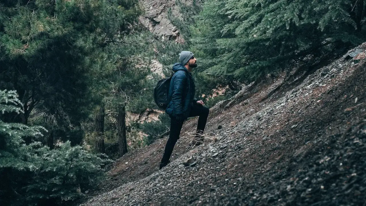 Randonneur en tenue d'hiver, sac &agrave; dos, escaladant un sentier rocailleux avec 100 m de d&eacute;nivel&eacute; positif dans une for&ecirc;t de pins.
