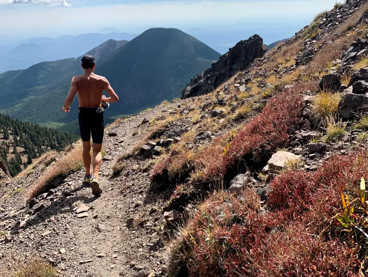 Un coureur sans torse d&eacute;vale un sentier fractionn&eacute; trail escarp&eacute;, entour&eacute; de v&eacute;g&eacute;tation rouge et de montagnes bleues &agrave; l'horizon.