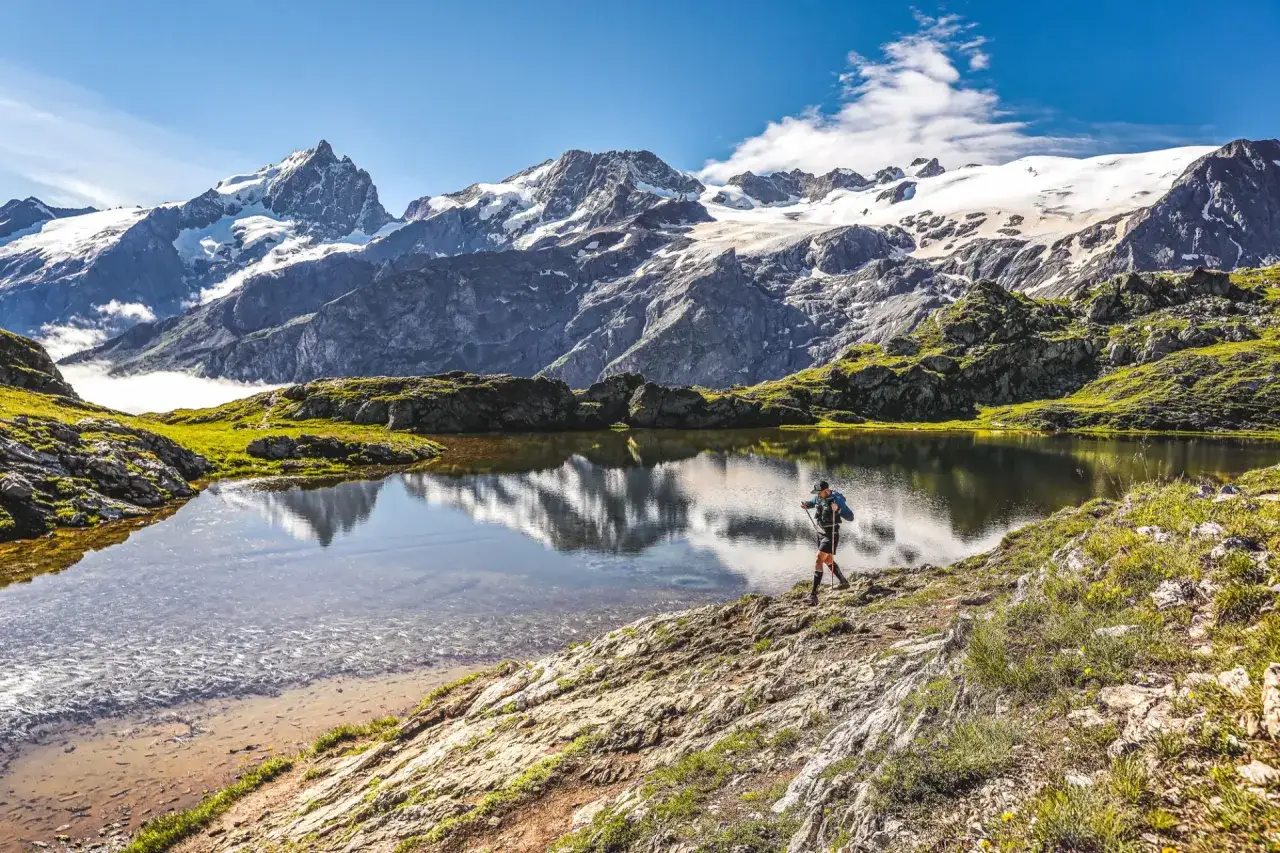 Un randonneur traverse le plateau d'Emparis, un lac alpin refl&eacute;tant des montagnes enneig&eacute;es sous un ciel bleu.
