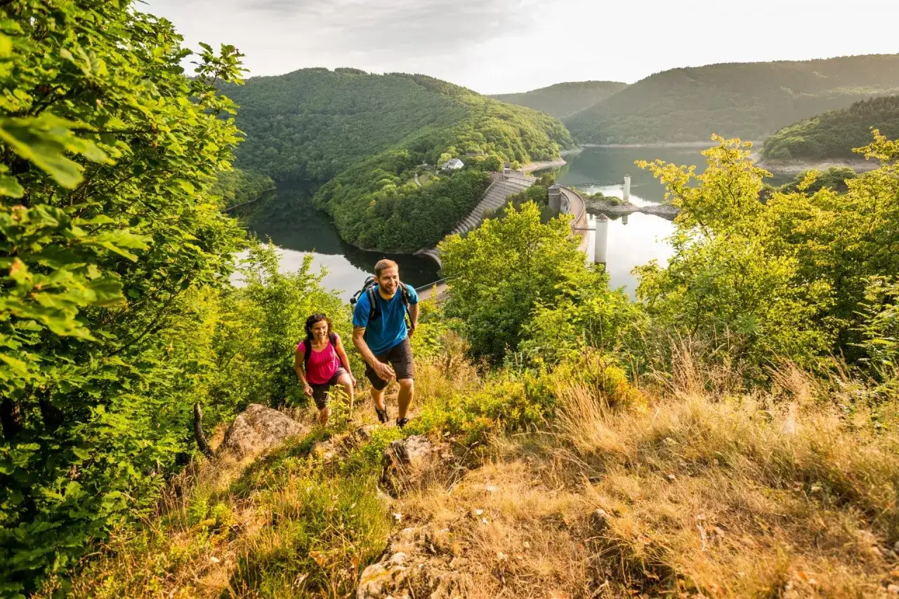 Couple randonnant sur un sentier verdoyant, avec un barrage et un lac en arri&egrave;re-plan, dans le parc national de l'Eifel.