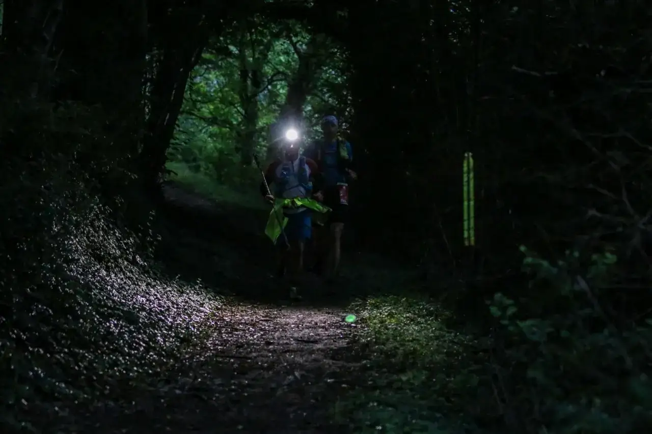 Deux coureurs s'aventurent dans un sentier forestier sombre, leurs lampes frontales éclairant le chemin. Un choix judicieux de lampe frontale est essentiel pour une telle course.
