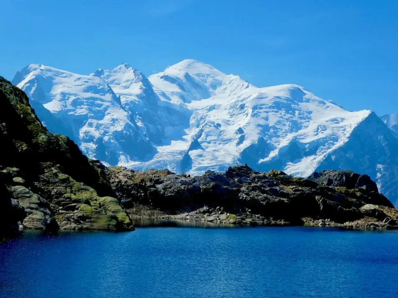 Lac d'altitude aux eaux bleues, reflétant les sommets enneigés du tour des Aiguilles Rouges sous un ciel clair.