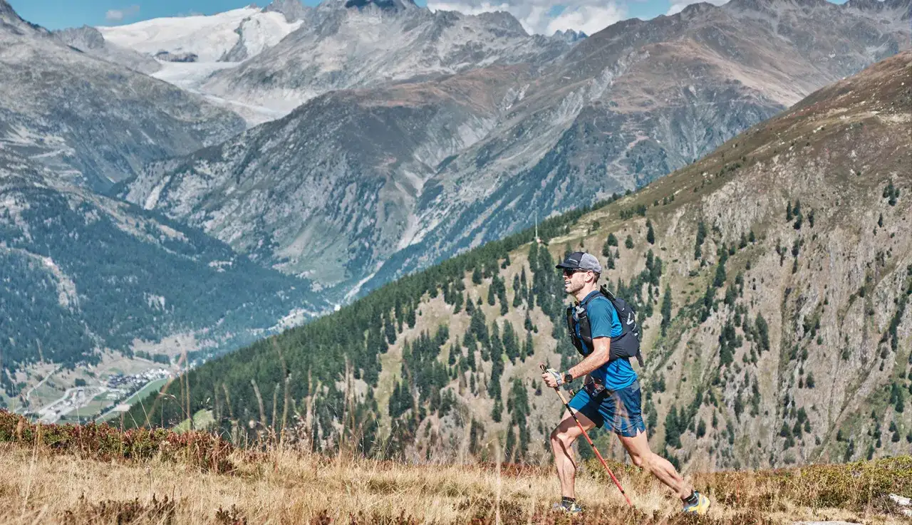 Un coureur en plein entra&icirc;nement en altitude, b&acirc;tons &agrave; la main, traverse un sentier de montagne escarp&eacute; avec des sommets enneig&eacute;s en arri&egrave;re-plan.