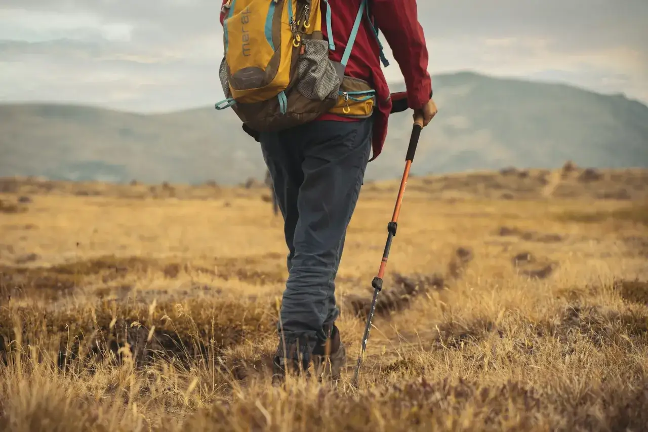 Randonneur en tenue de sport, sac &agrave; dos jaune et rouge, utilisant un b&acirc;ton de marche pliable ou t&eacute;lescopique pour gravir une colline herbeuse.