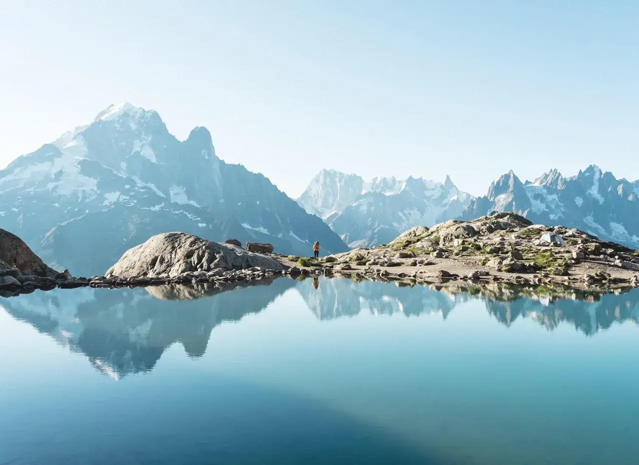Une personne profite des randonn&eacute;es en France, au bord d'un lac alpin refl&eacute;tant des montagnes enneig&eacute;es.